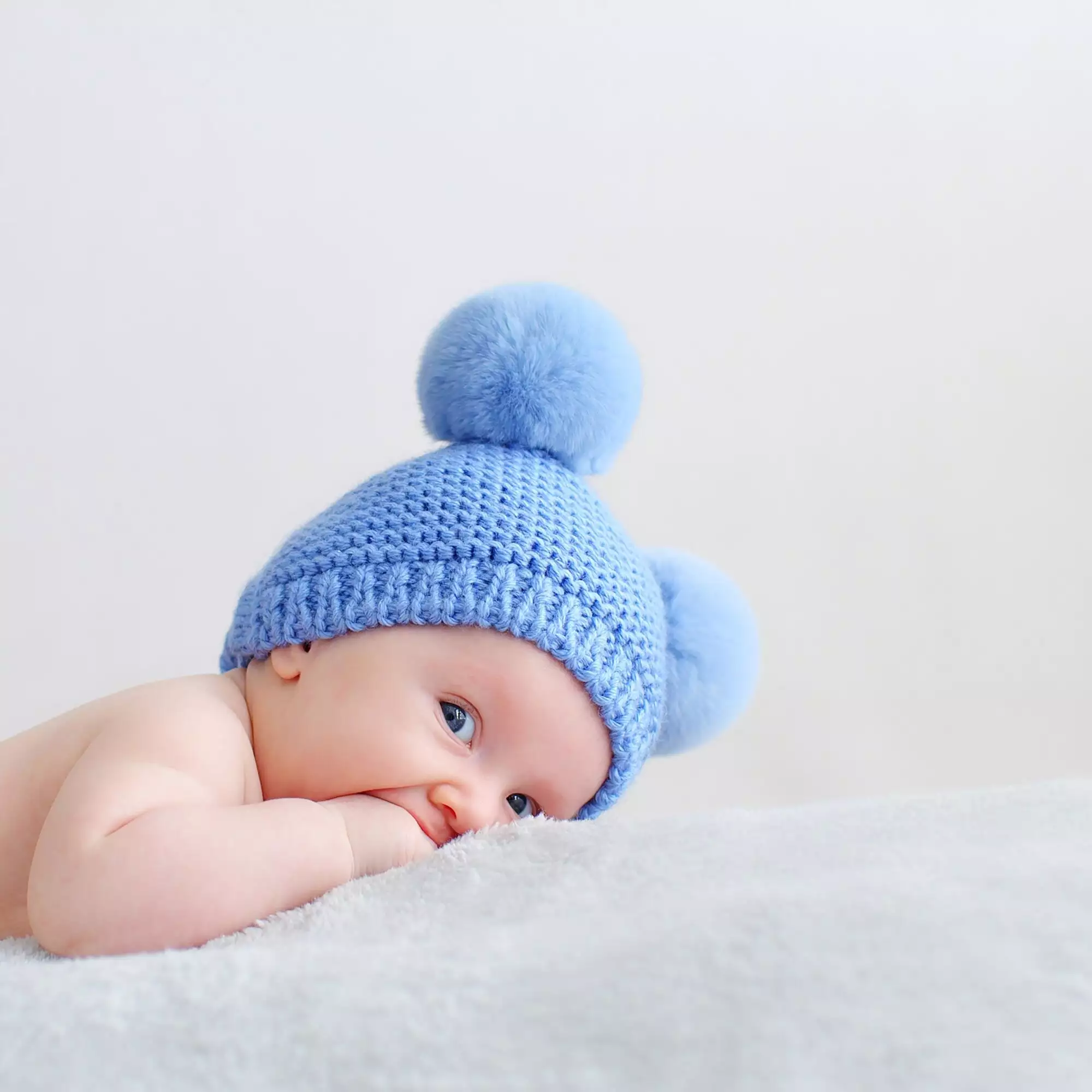 Sweet newborn baby in a blue hat with pompoms looks into the camera.